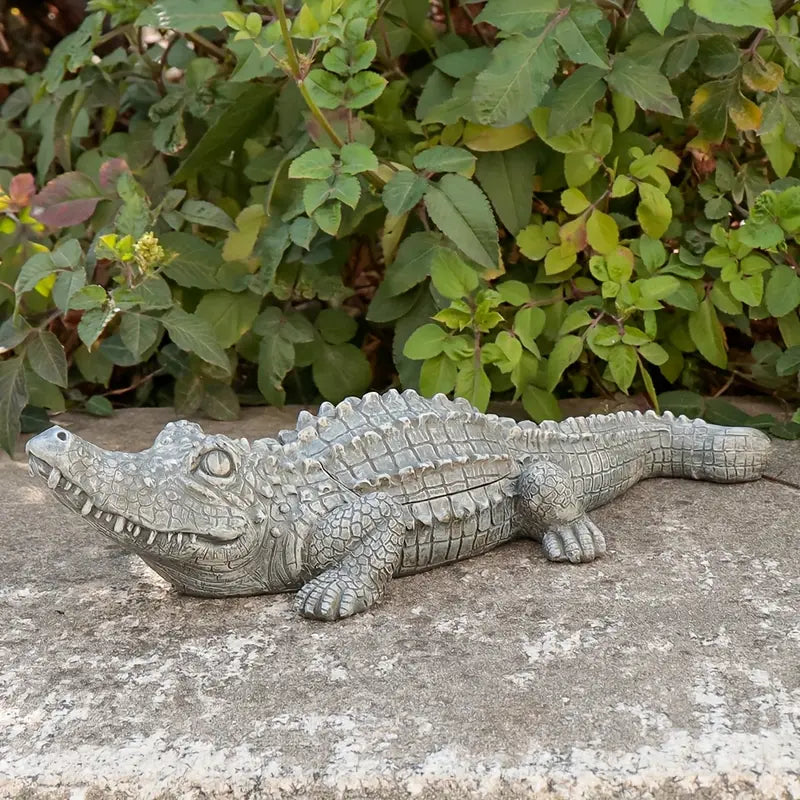 Gray alligator statue on a concrete surface with green foliage in the background